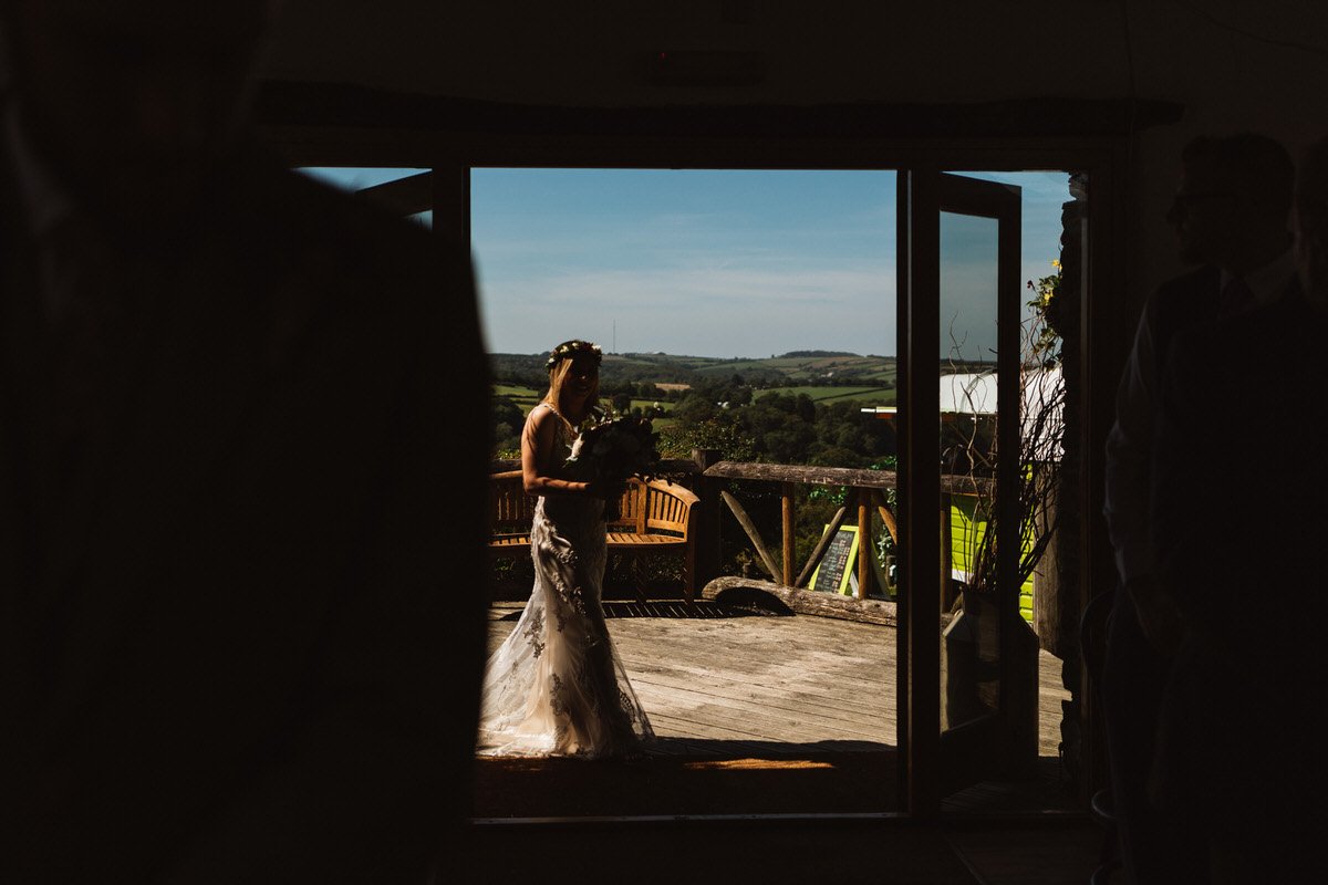 Bride entering the ceremony overlooking a valley. UK wedding photographer captures her half in light and half silhouette